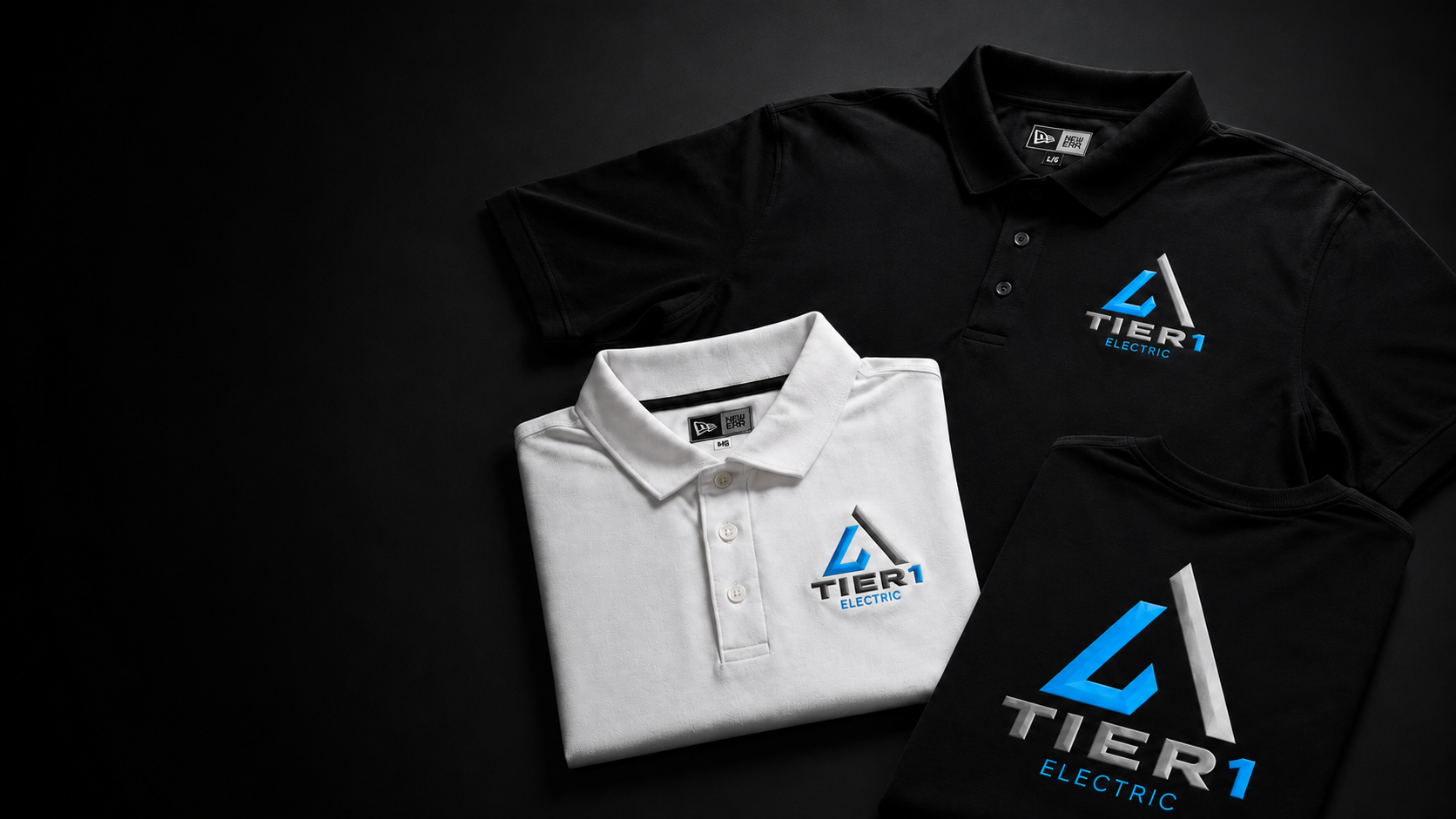 Three folded polo shirts and a folded T-shirt displayed on a dark background, each featuring the same blue and white 'TIER 1 ELECTRIC' logo. One shirt is white, while the others are black.
