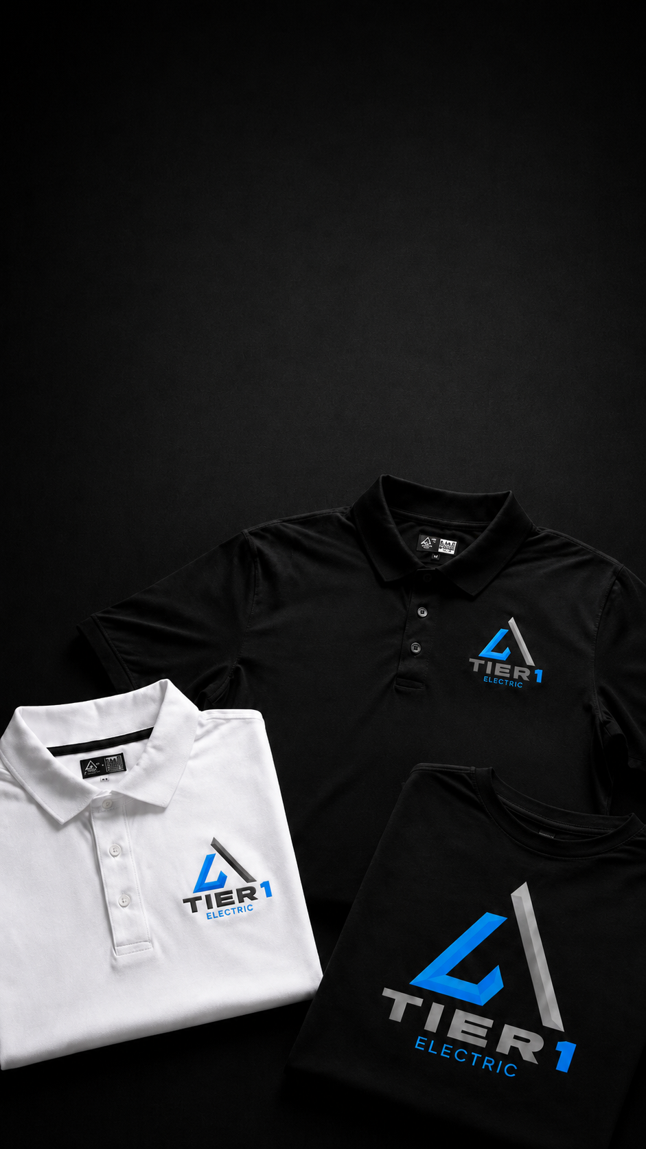 Two folded polo shirts and a folded T-shirt displayed on a dark background. The black shirt at back right has a blue and white Tier 1 Electric logo on the left chest, the white shirt at lower left has the same logo on the left chest, and the black shirt at lower right has a large blue and white Tier 1 Electric logo on the front.