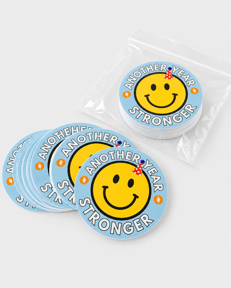 Stack of custom Another Year Stronger smiley face stickers with blue background, birthday hat graphic, and clear packaging.